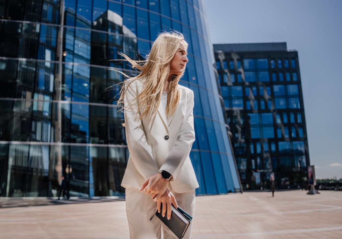Junge blonde Frau im weißen Anzug steht draußen vor einem gläsernen Gebäude.