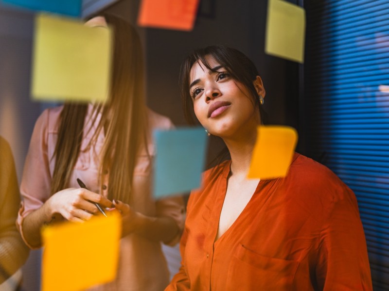 Geschäftsfrau in einem orangefarbenen Hemd liest während einer Brainstorming-Sitzung nachdenklich Notizen an einer Glaswand.