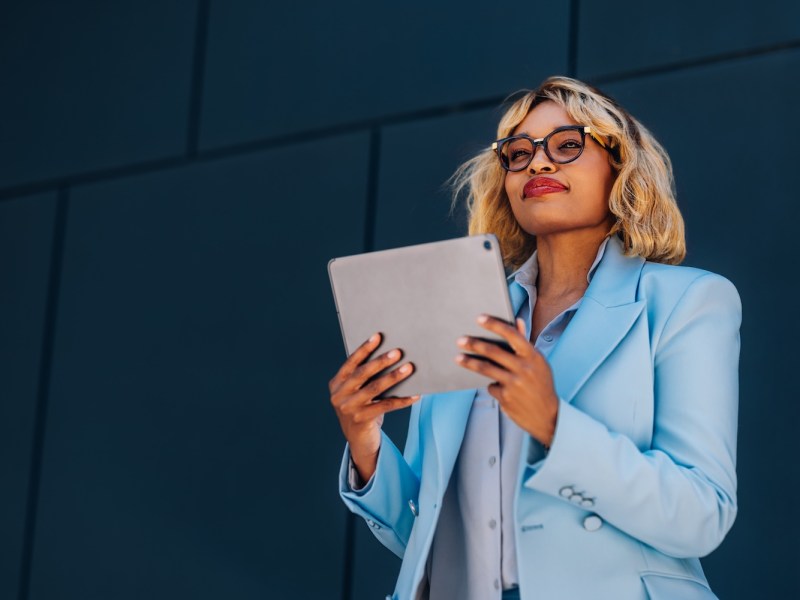 Selbstbewusste Geschäftsfrau mit Brille und blauem Blazer hält ein Tablet vor einem Bürogebäude und blickt nach oben.
