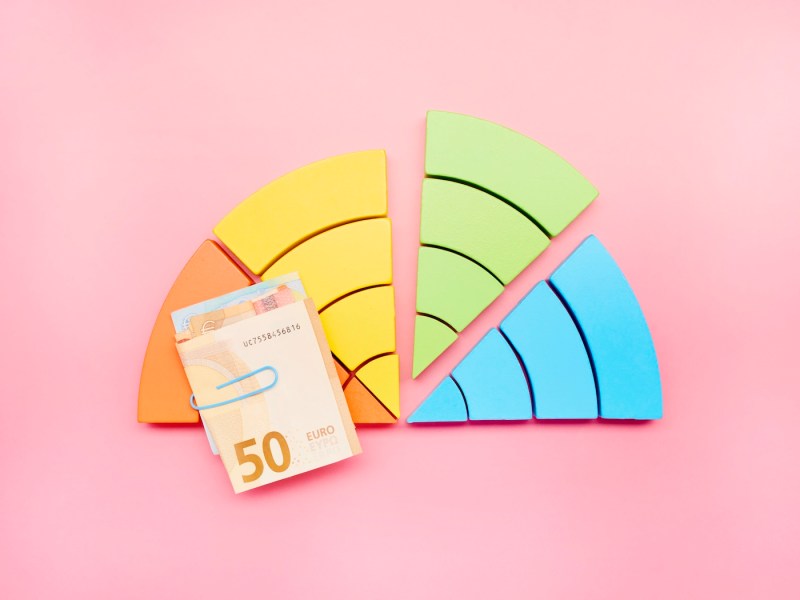 Ein halbes Kreisdiagramm aus bunten Bausteinen und einem Haufen Euro-Banknoten auf rosa Hintergrund, das grüne Viertel fällt auf.