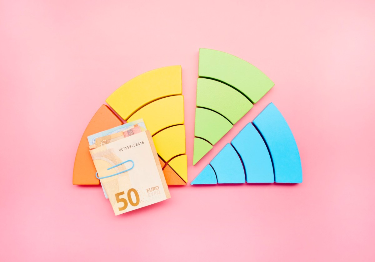 Ein halbes Kreisdiagramm aus bunten Bausteinen und einem Haufen Euro-Banknoten auf rosa Hintergrund, das grüne Viertel fällt auf.