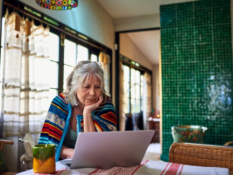 Ältere Frau, in eine Decke gehüllt, steht am Esstisch und schaut sich E-Mails auf dem Laptop an.