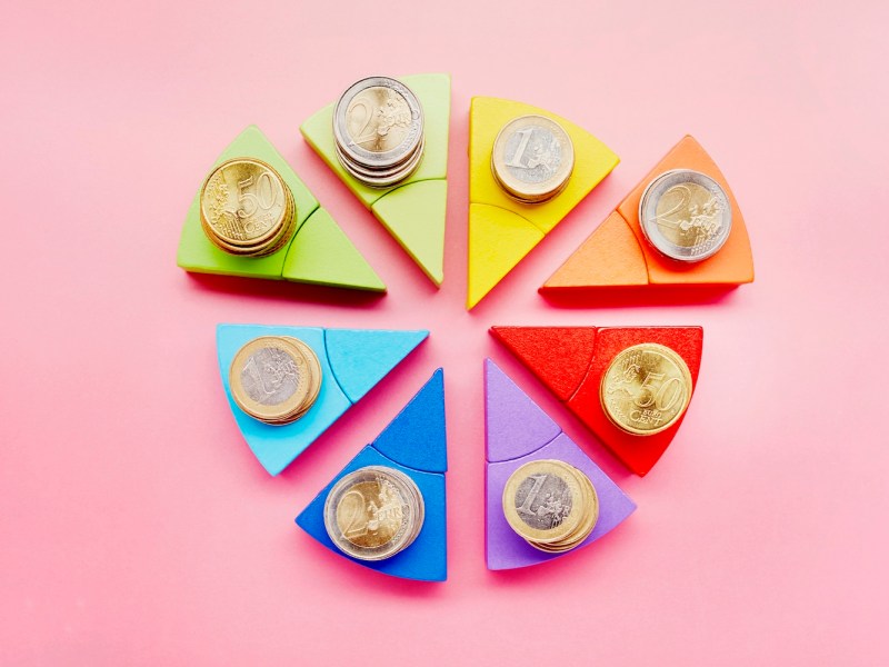 Kreisdiagramm aus bunten Bausteinen und Stapeln von Euro-Münzen auf rosa Hintergrund.