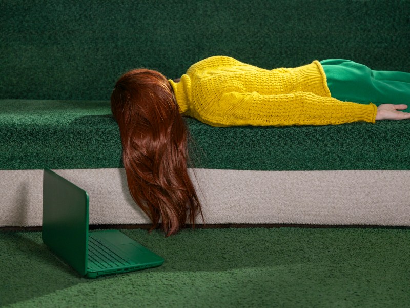 Rothaarige Frau liegt zu Hause mit einem Laptop auf einer Couch und wirkt müde und ausgeruht.