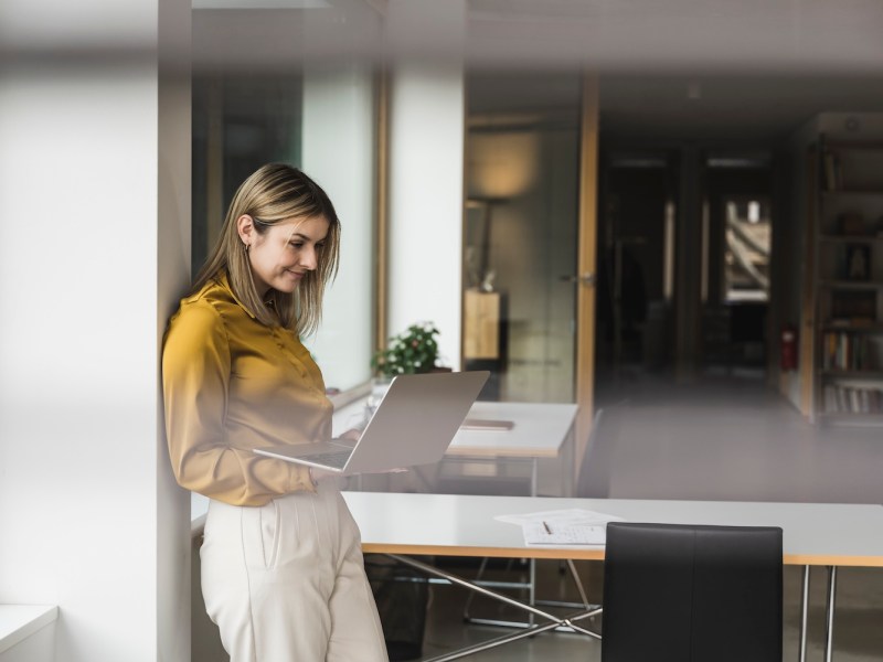 Lächelnde Geschäftsfrau lehnt an der Wand und arbeitet am Laptop im Büro.