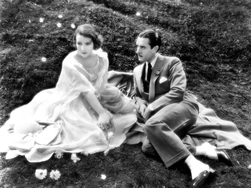 Frau und Mann sitzen im Kleid und Anzug auf der Wiese in Schwarz-Weiß-Foto.