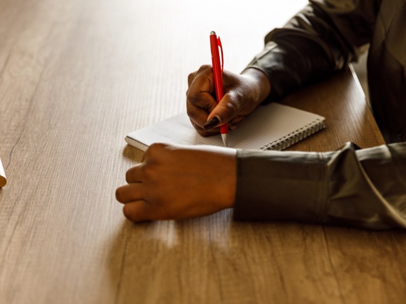 Jungen schwarze Frau, die am Schreibtisch ihres Homeoffice sitzt und in einen Notizblock eine To-do-Liste für den Tag schreibt.