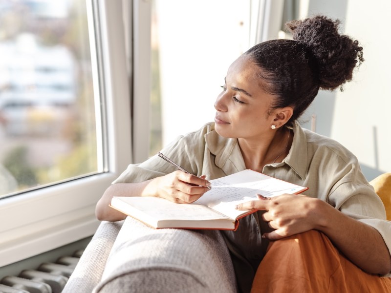 Eine junge Frau ist zu Hause, sie sitzt auf dem Sofa im Wohnzimmer und macht sich Notizen.