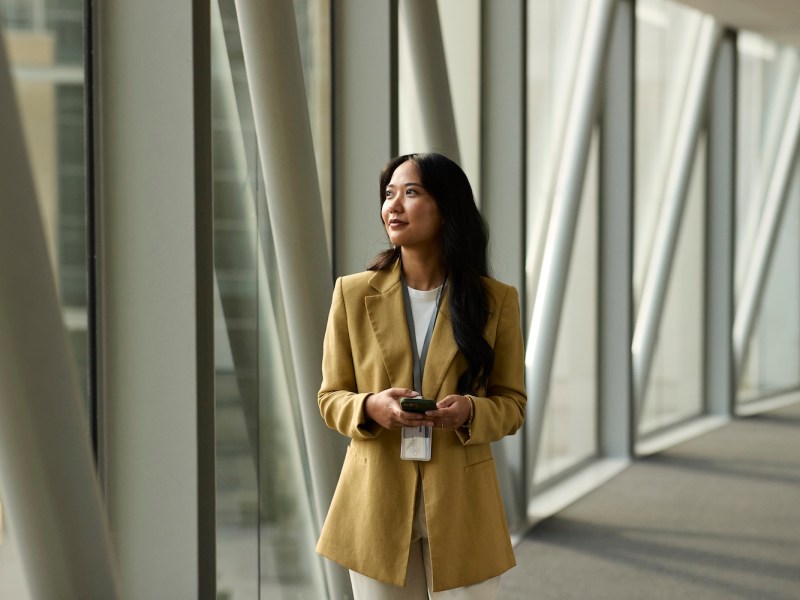 Eine Business-Frau hält ihr Smartphone in der Hand und schaut durch eine Glaswand.