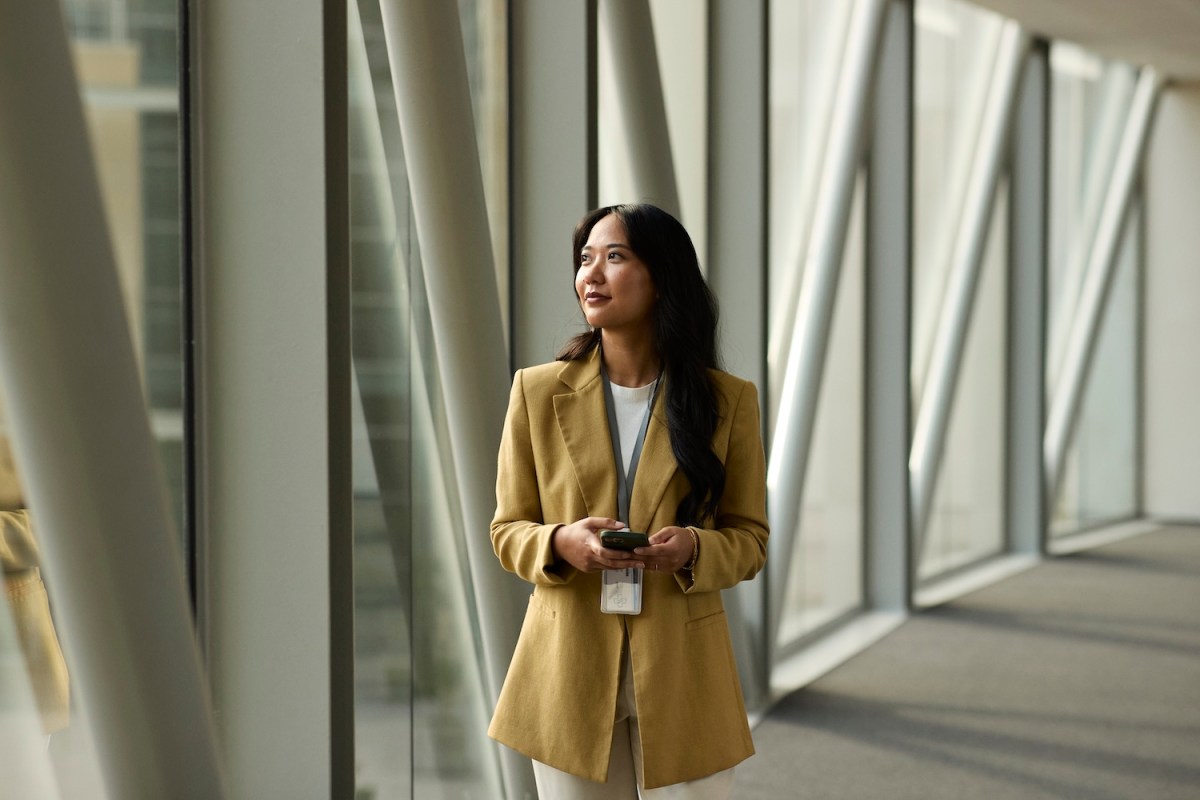 Eine Business-Frau hält ihr Smartphone in der Hand und schaut durch eine Glaswand.