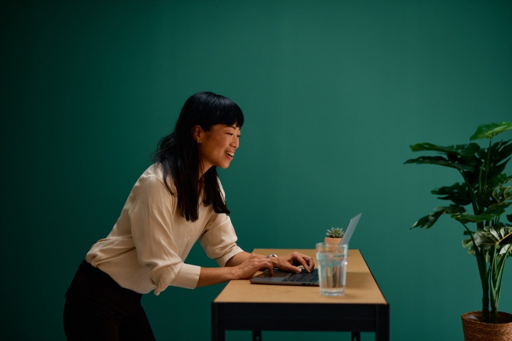 Junge asiatische Gesch&auml;ftsfrau lehnt &uuml;ber einem Schreibtisch, tippt auf einem Laptop und l&auml;chelt.