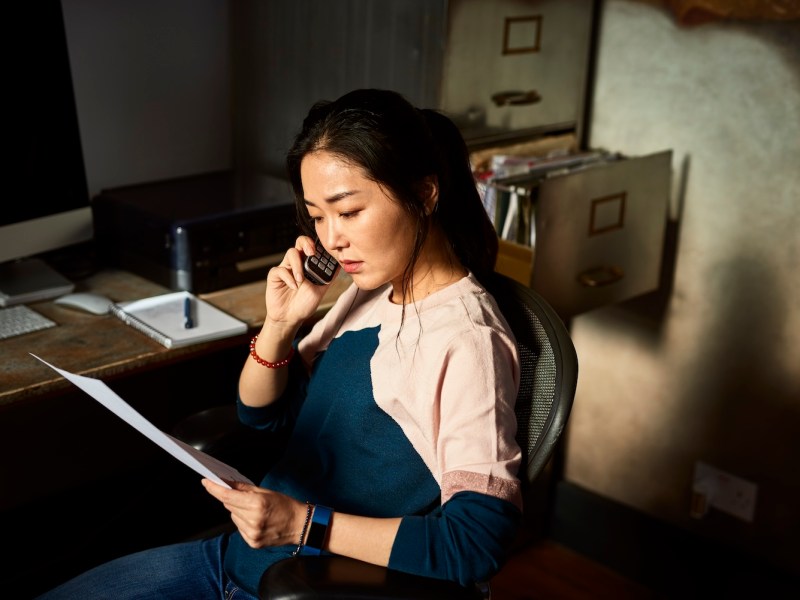 Asiatische Frau mittleren Alters, die am Telefon zuhört, im Bürostuhl in einem dunklen Raum sitzt, Papierkram hält.