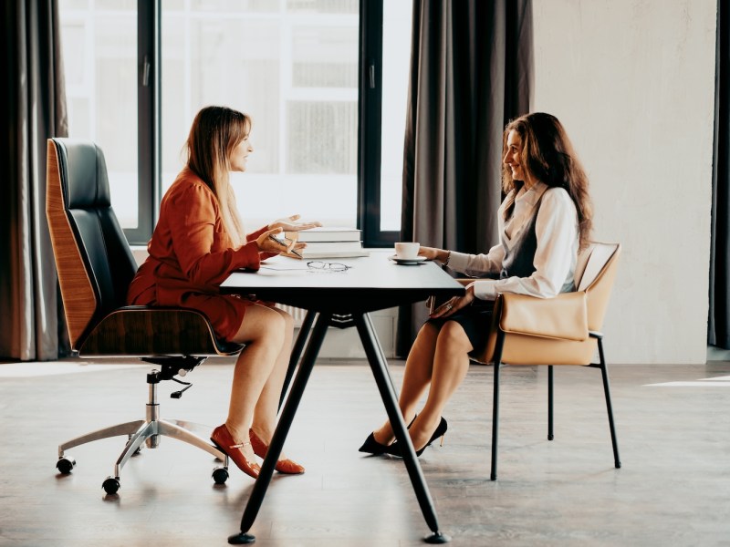 Zwei Geschäftsfrauen sitzen sich in einem modernen, hell erleuchteten Büro gegenüber und unterhalten sich.