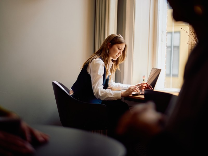 Eine konzentrierte Geschäftsfrau sitzt an einem Fenster und tippt auf ihrem Laptop an einem Holzschreibtisch.