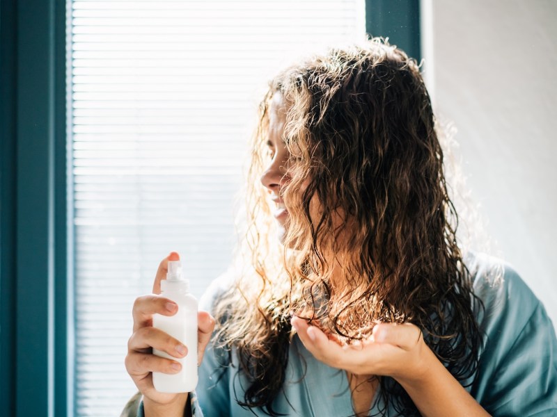 Eine Frau mit nassem, gewelltem Haar trägt ein Pflegeprodukt aus einer Sprühflasche auf.