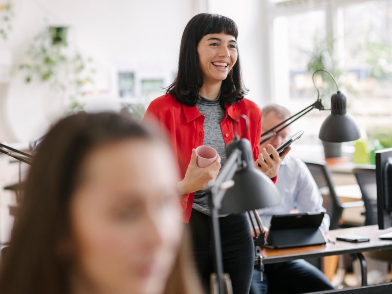 Eine lächelnde Geschäftsfrau, die mit einem Laptop und einer Kaffeetasse in der Hand durch ihr Büro geht.
