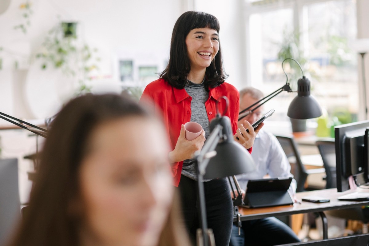 Eine lächelnde Geschäftsfrau, die mit einem Laptop und einer Kaffeetasse in der Hand durch ihr Büro geht.