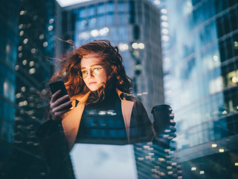 Frau nutzt nachts im Freien ihr Smartphone in der Nähe eines Wolkenkratzers.