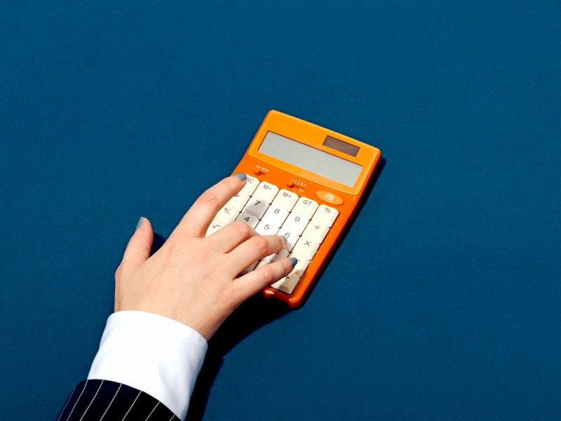 Nahaufnahme einer Hand, die einen orangefarbenen Retro-Taschenrechner vor blauem Hintergrund benutzt.