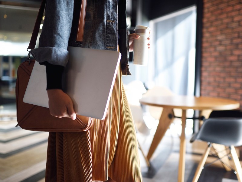 Eine junge Frau hält einen Laptop unter dem Arm und in der anderen Hand hält sie eine Flasche.